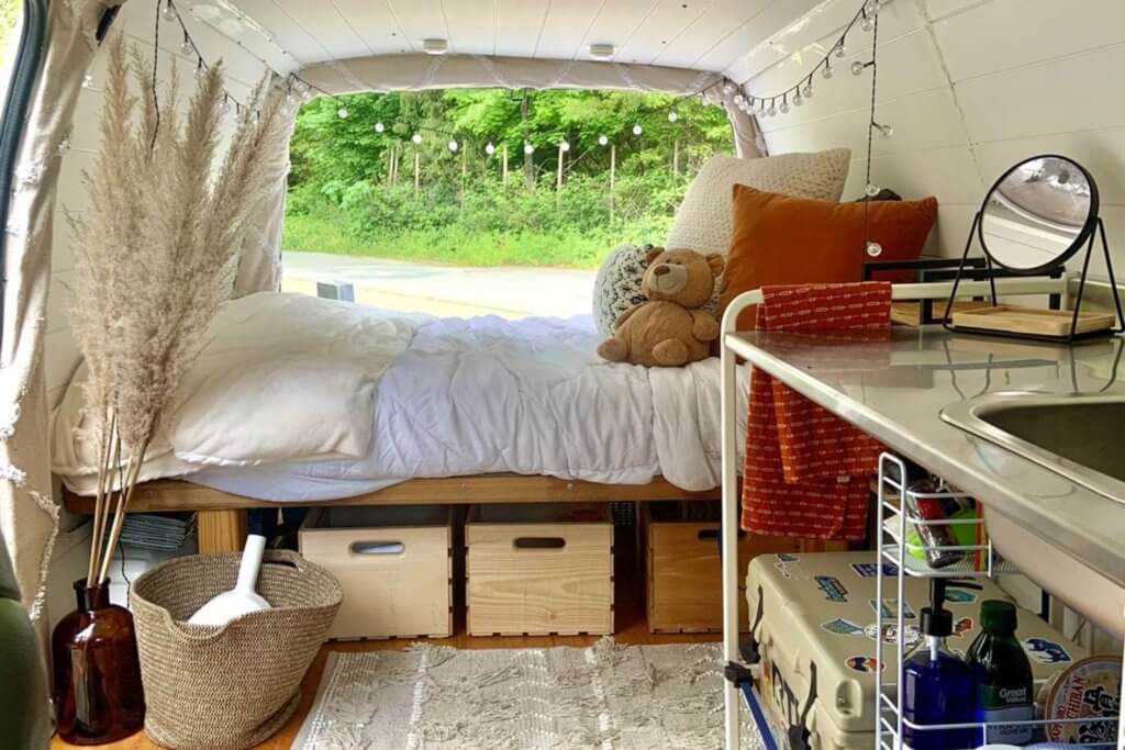 Boho bed camper van style. White sheets and orange pillow with teddy bear pillow.